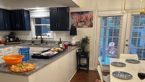 Ett kök med mörka skåp, en bänkskiva i granit, en matplats med ett vitt bord och blå tallrikar samt ett fönster med persienner.