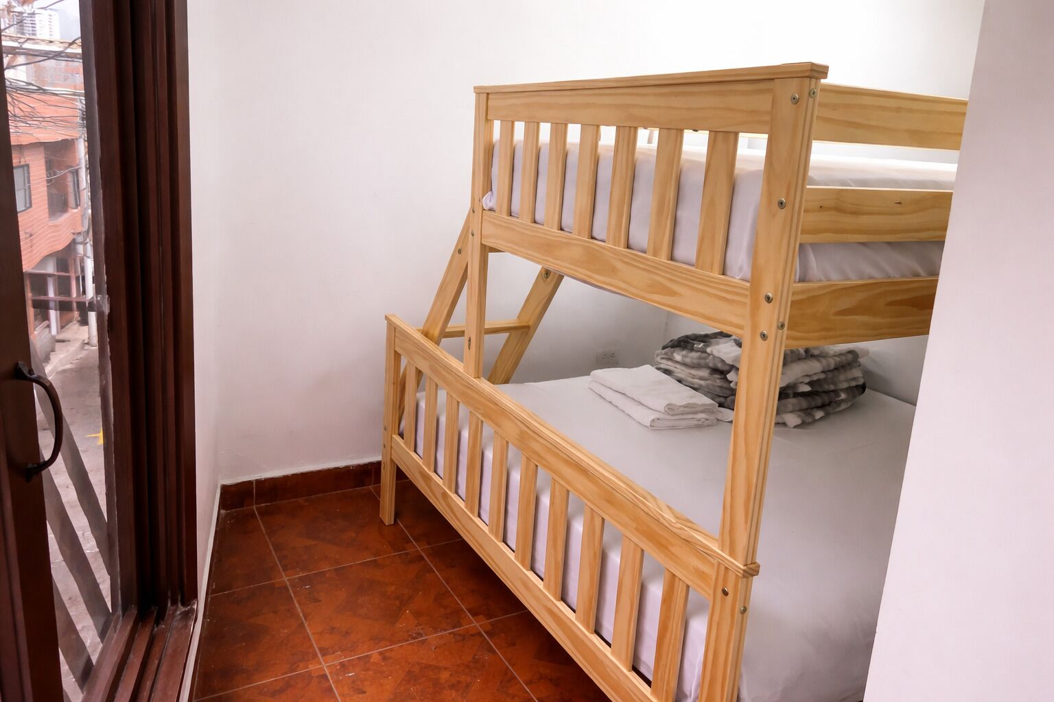 Een stapelbed met houten frame en witte beddengoed, een betegelde vloer en uitzicht op een gebouw door een deur.