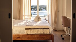A bedroom with a bed, a chair, and a view of the city through large windows. - Outsite Vilamoura - Work, Connect, Explore (Loule)