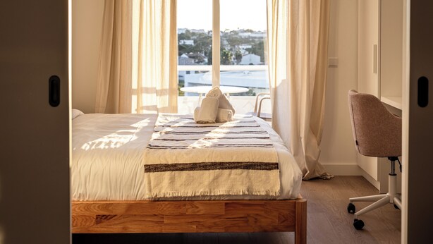 A bedroom with a bed, a chair, and a view of the city through large windows. - Outsite Vilamoura - Work, Connect, Explore (Loule)