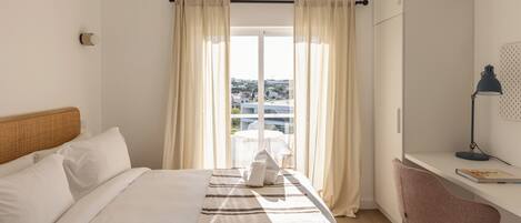 A hotel room with a bed, a desk, a chair, a lamp, and a view through the window.