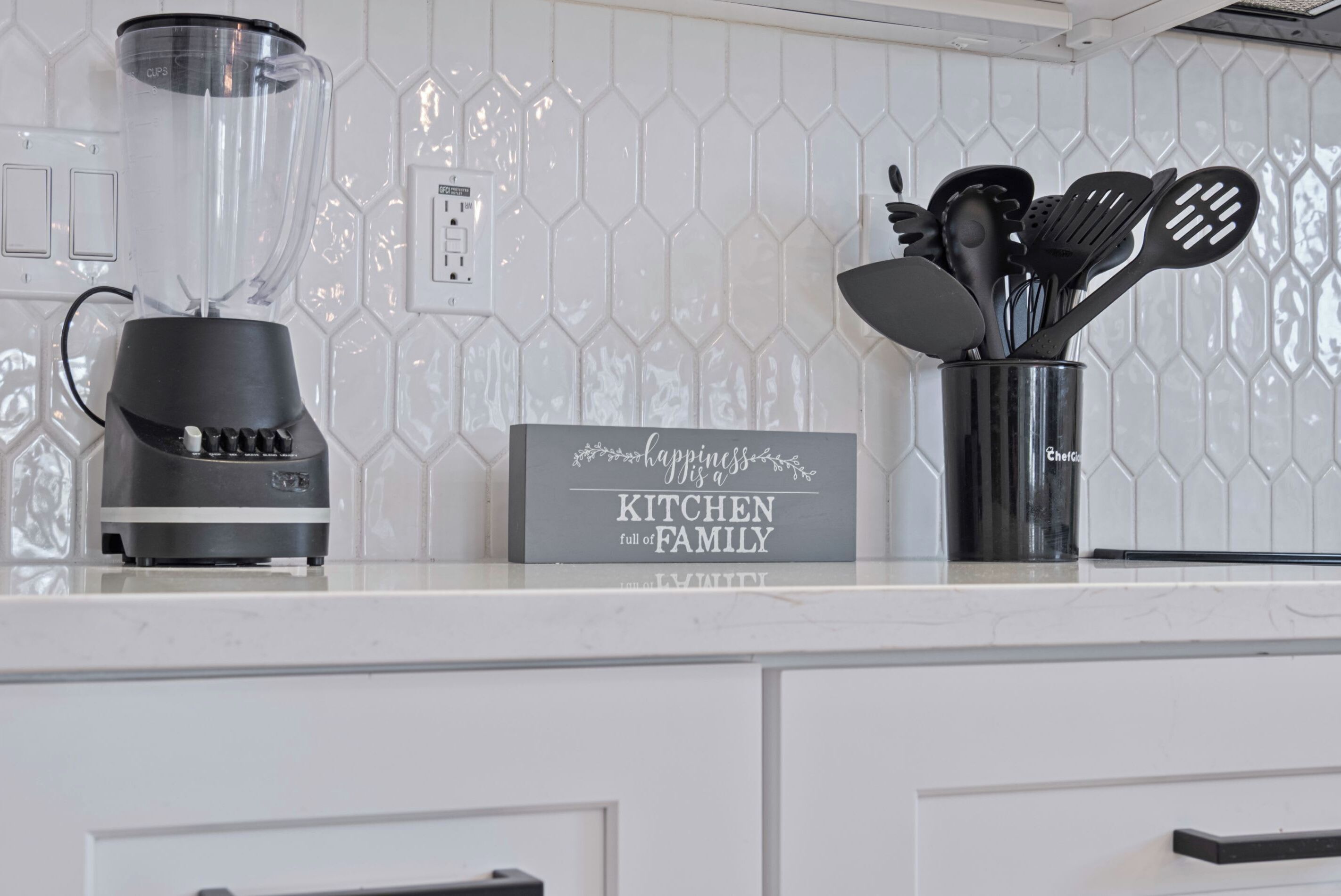 A kitchen counter with a blender, a sign that says "happiness is a kitchen full of family," and a utensil holder.