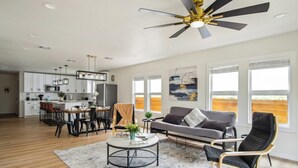 A spacious living area with a dining table, sofa, and chairs, a ceiling fan, and large windows.