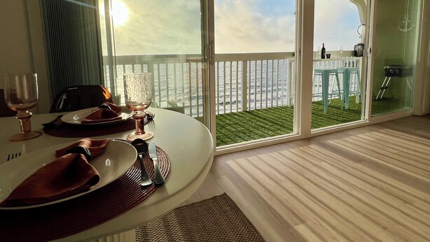 A dining area with a table set for two, large windows with a view of the sea, and a balcony with a white railing. - Oceanfront Escape - Steps to Beach - Stunning Views & Balcony (Redondo Beach)