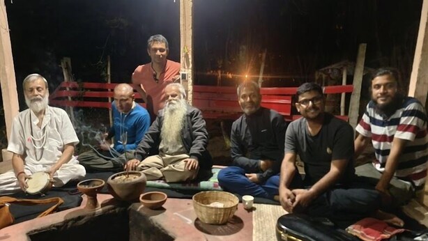 A group of people sitting around a traditional setup with a fire pit, bowls, and a mat. - Golok Riverside Retreat (Bolpur)