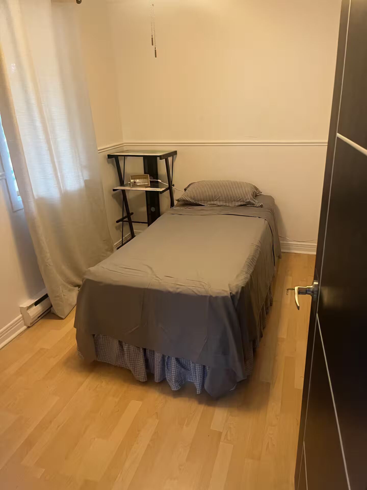 A small bedroom with a bed, a nightstand, a window with curtains, and a door.
