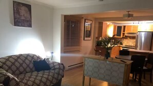 Ein Wohnzimmer mit einem gemusterten Sofa, einem kleinen Tisch und einer Küchenzeile inklusive Kühlschrank und Schränken.