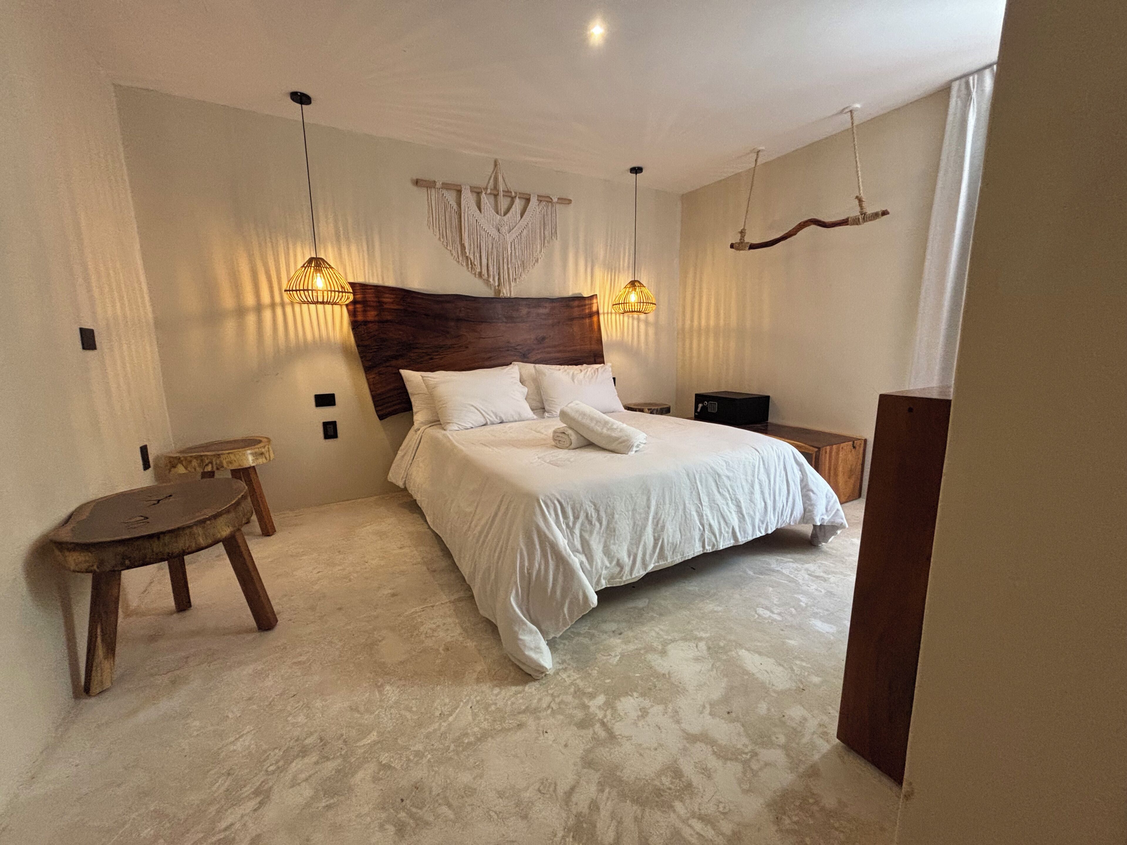 A bedroom with a large bed, two bedside tables, a small stool, a macrame wall hanging, and two hanging light fixtures.