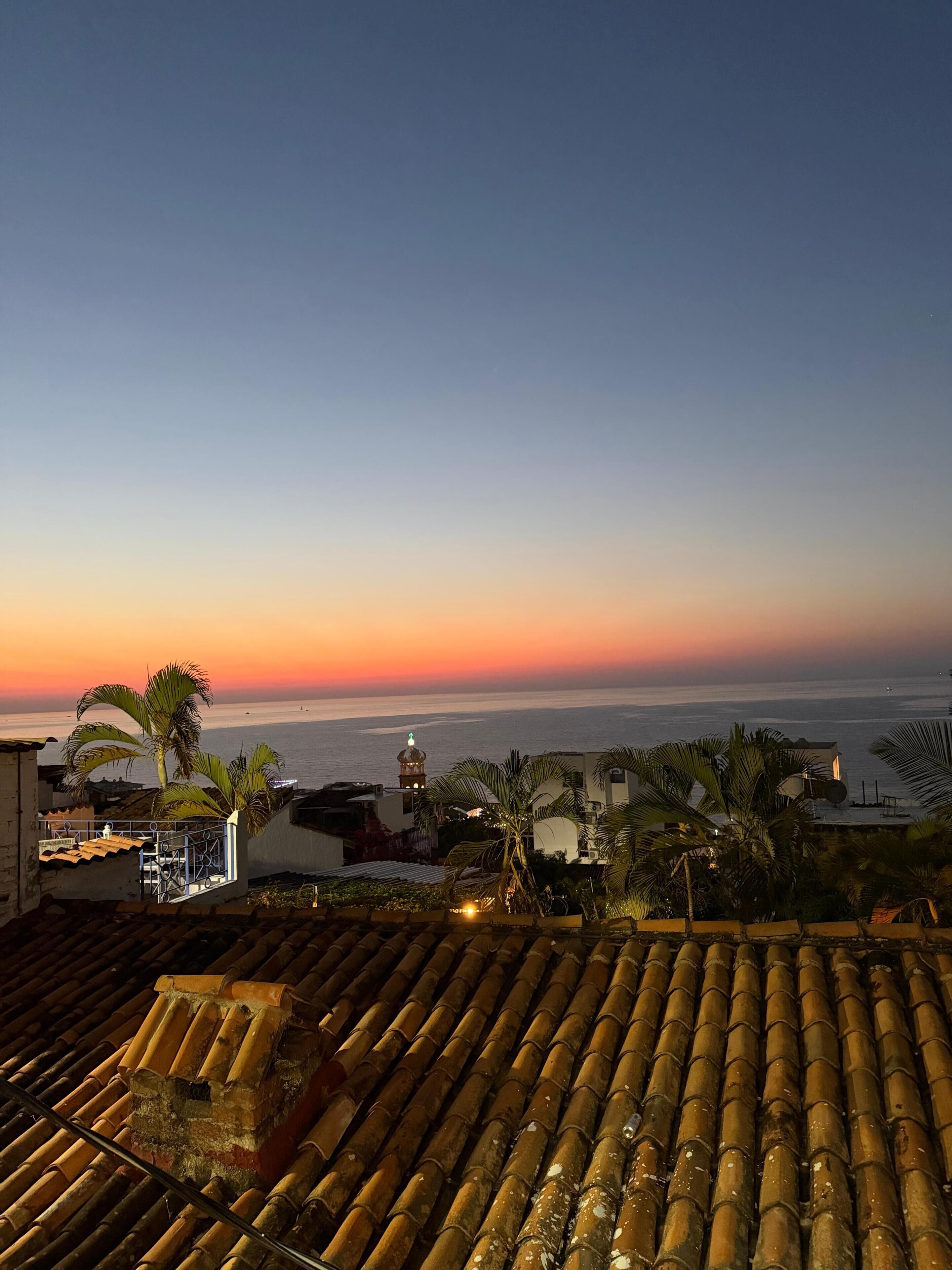 A coastal sunset view from a rooftop with a tiled roof, palm trees, and a distant building with a dome.