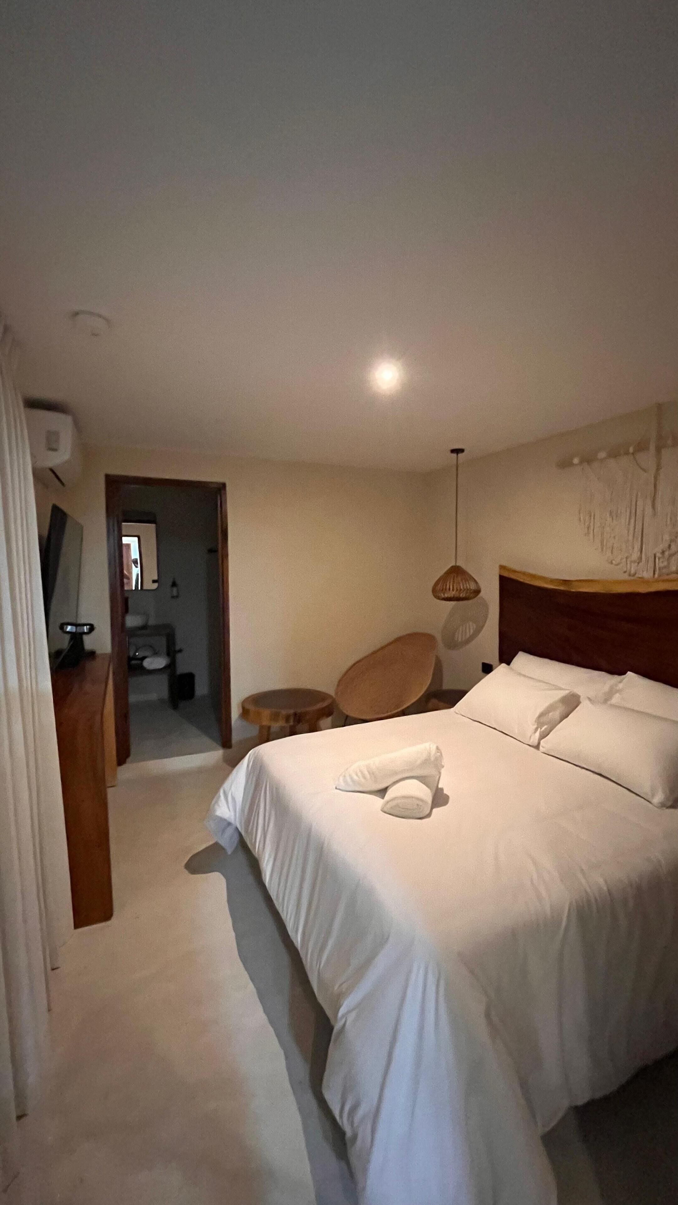 Una habitación de hotel con una cama grande, cabecera de madera, mesita de noche, una silla y una lámpara colgante.