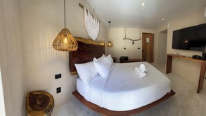 A bedroom with a curved bed, a wooden headboard, a hanging macrame wall decoration, a television, and a wooden side table with a kettle. - Hotel Boutique Alcalá Vallarta (Puerto Vallarta)