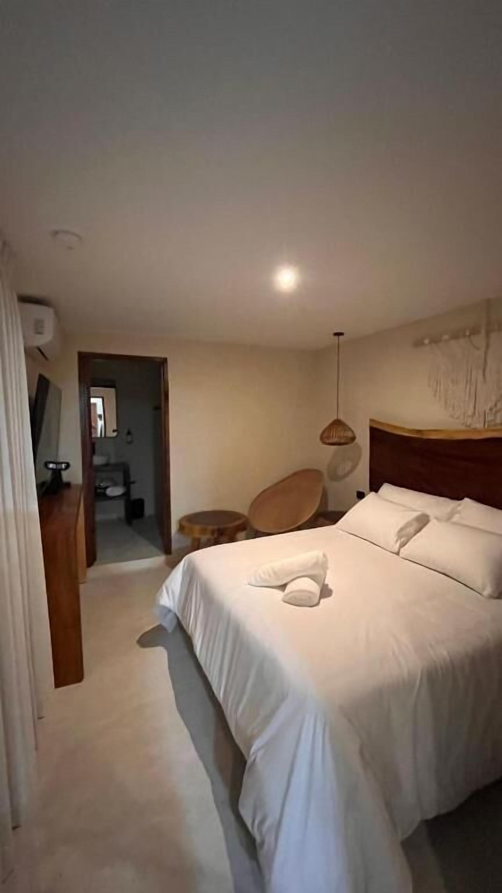 Une chambre d’hôtel avec un grand lit, une tête de lit en bois, une table de chevet, une lampe suspendue et une vue sur la salle de bain.