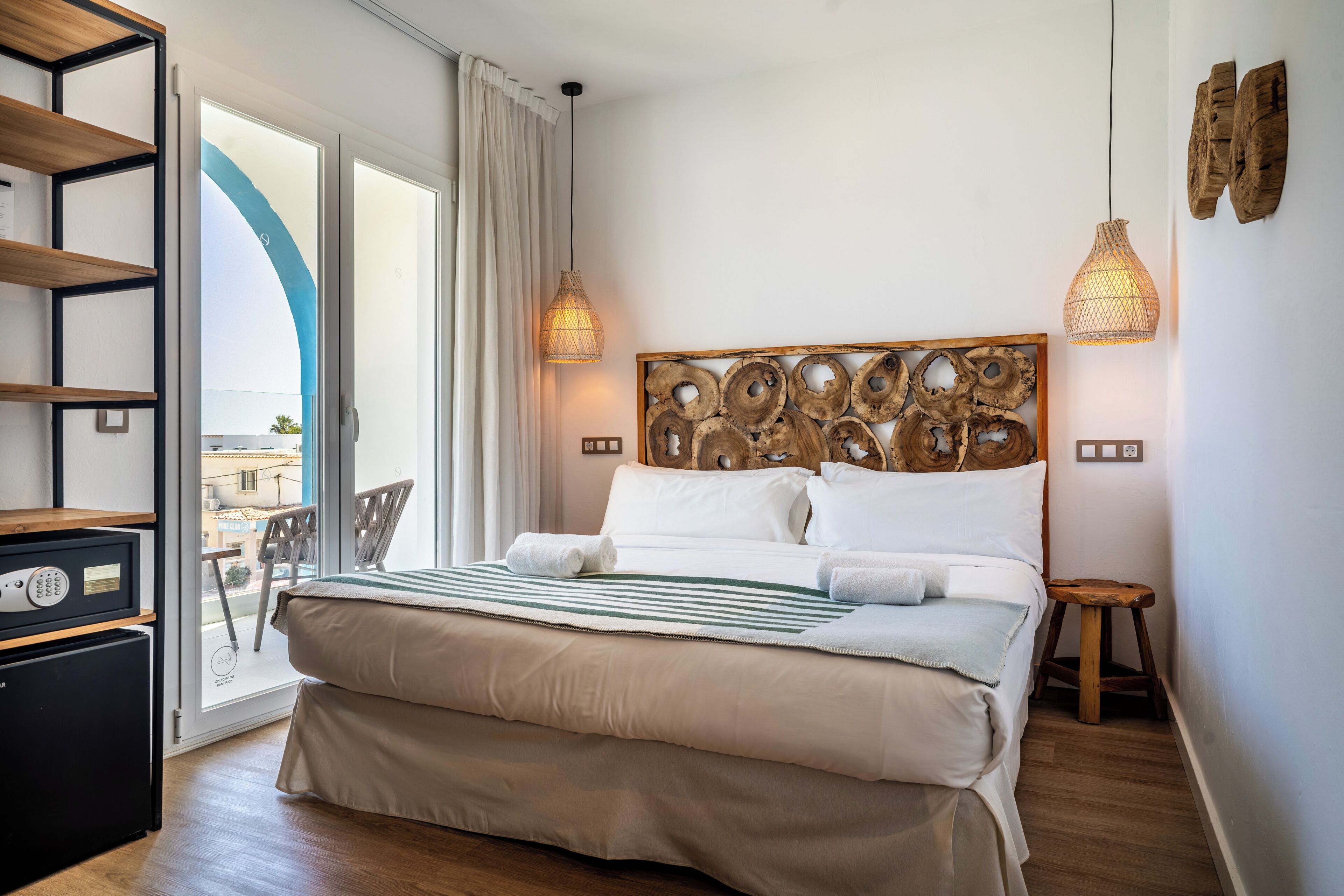 Habitación de hotel con cama, escritorio, silla, taburete pequeño, estante en la pared y vistas a través de una puerta de cristal.