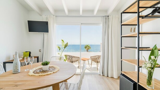 A modern hotel room with a wooden dining table, a TV mounted on the wall, a balcony with a view of the sea, and a built-in bookshelf. - Outsite Ibiza - Work, Connect, Explore (Santa Eulalia del Rio)