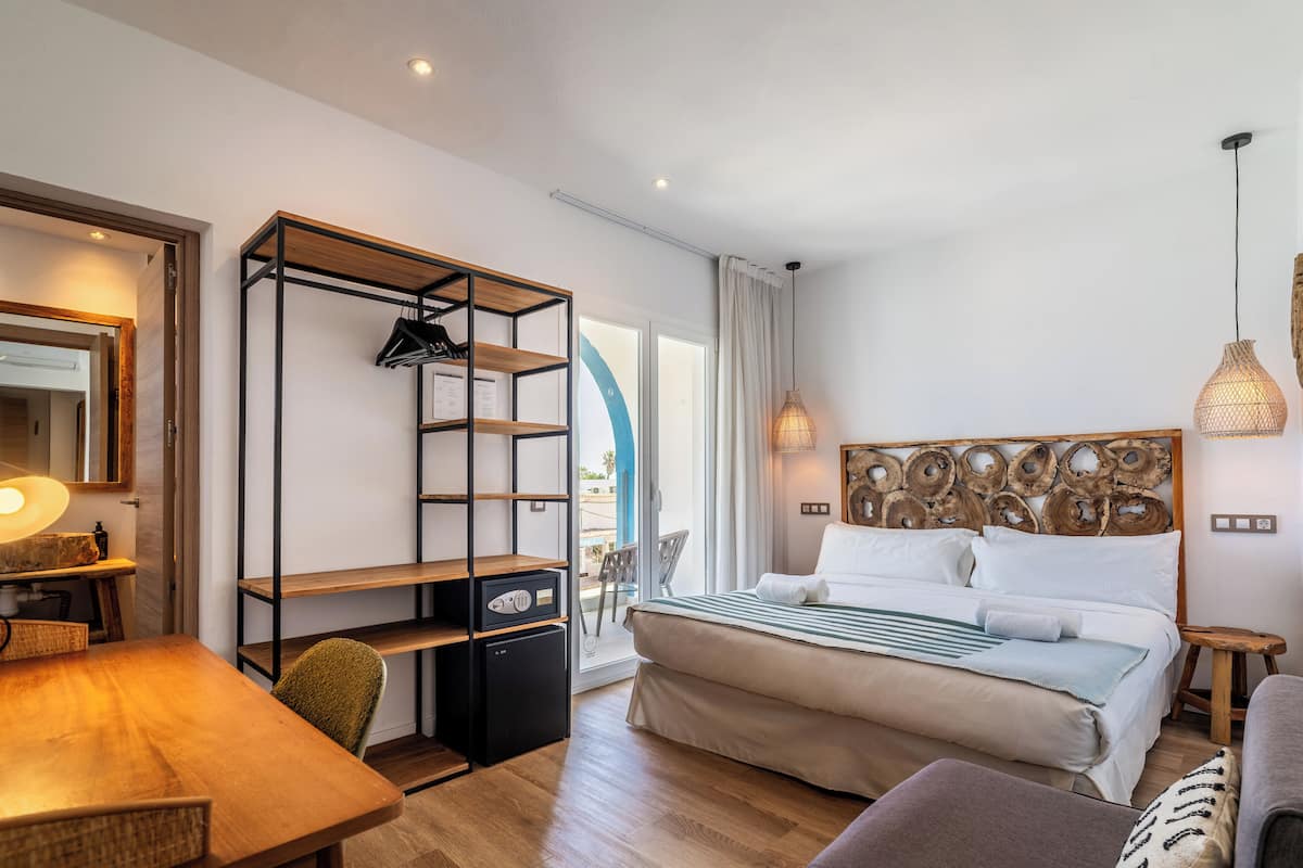 Una habitación de hotel moderna con un escritorio de madera, una cama con cabecera, un pequeño rincón de descanso y un balcón con vistas.