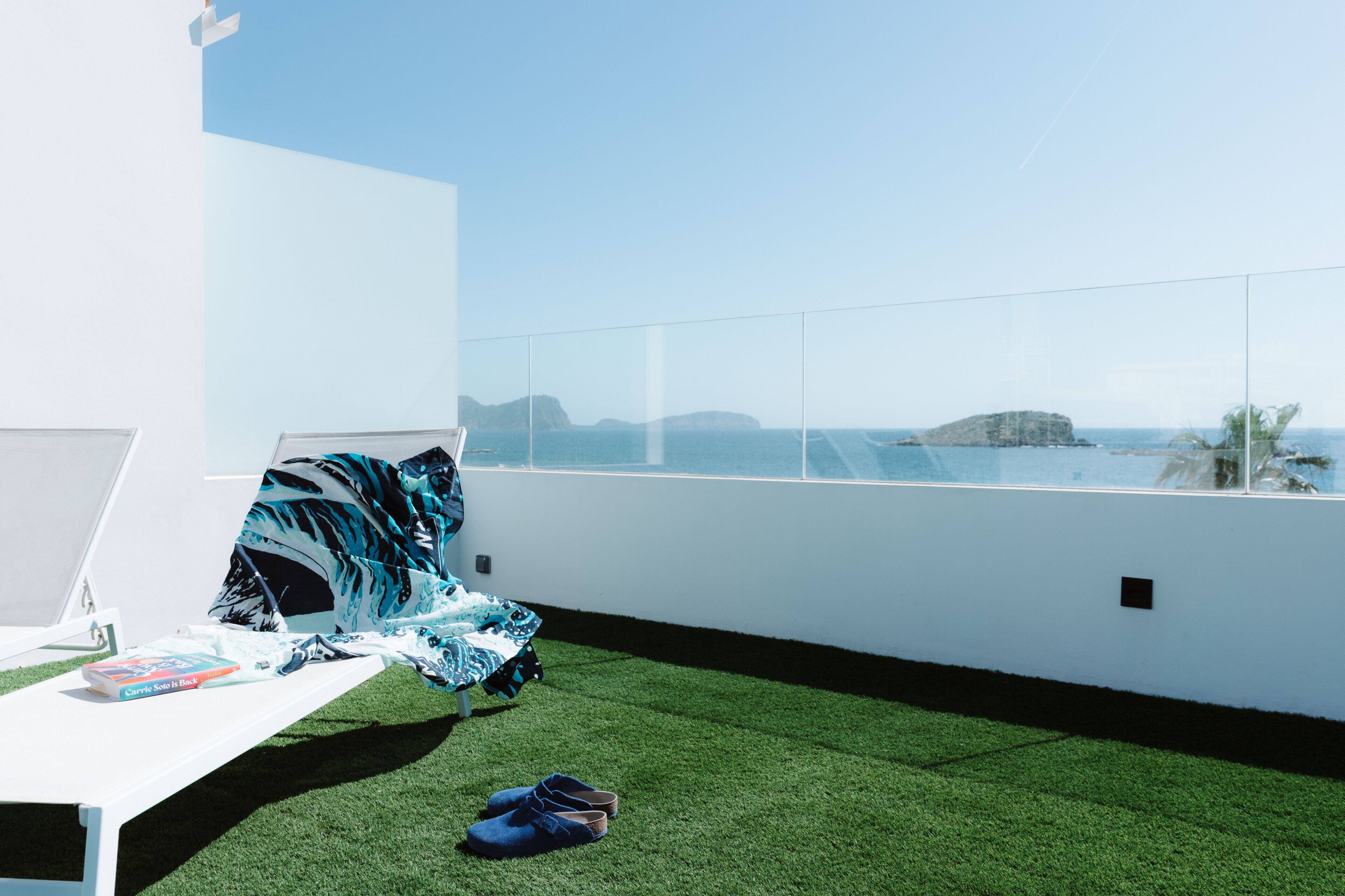 A room with a white wall, a bed with a blue and white patterned blanket, a pair of shoes on the floor, and a view of the sea and mountains.