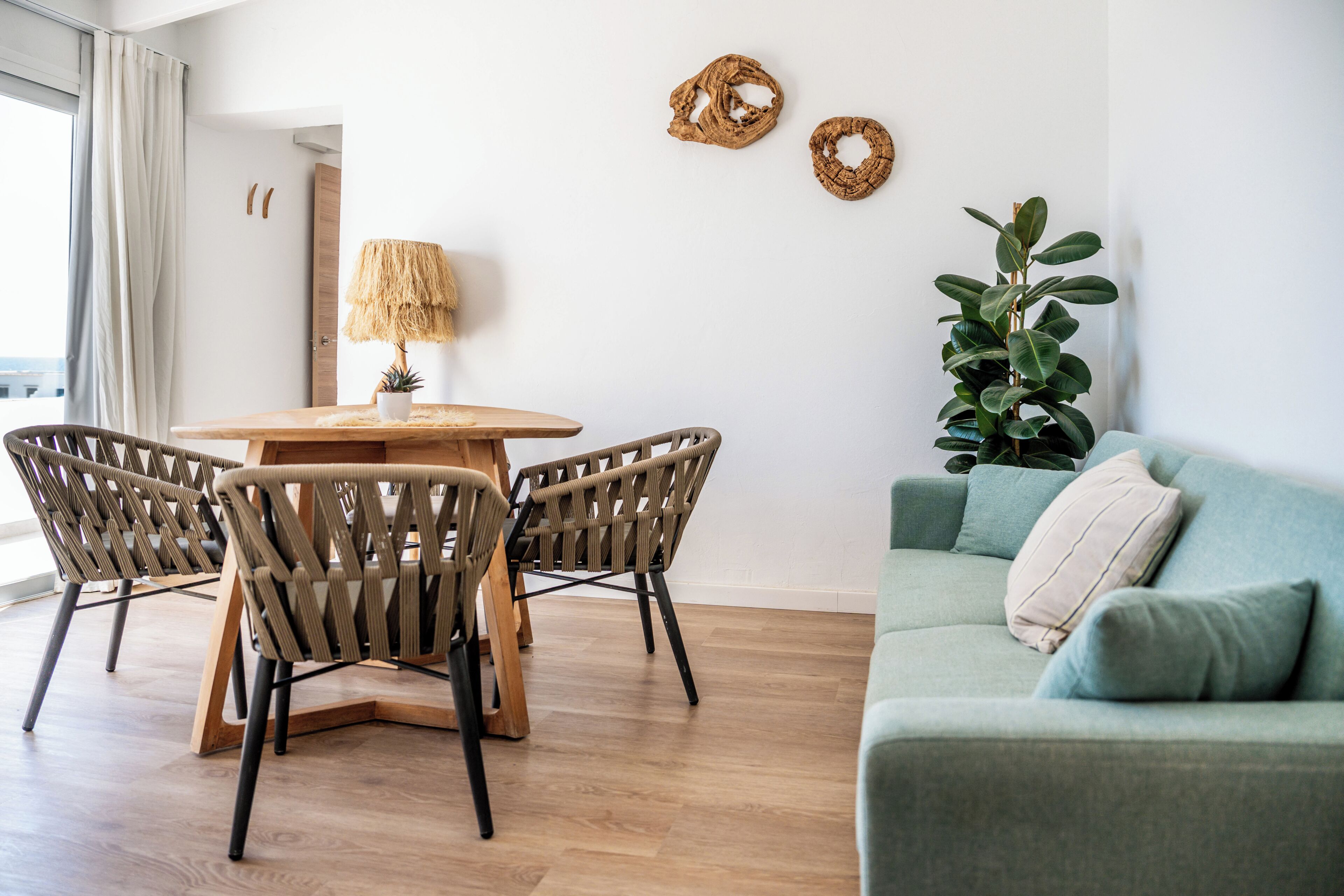 Ein Zimmer mit einem Holztisch, um den herum Korbstühle stehen, einem grünen Sofa mit Kissen und einer Zimmerpflanze.