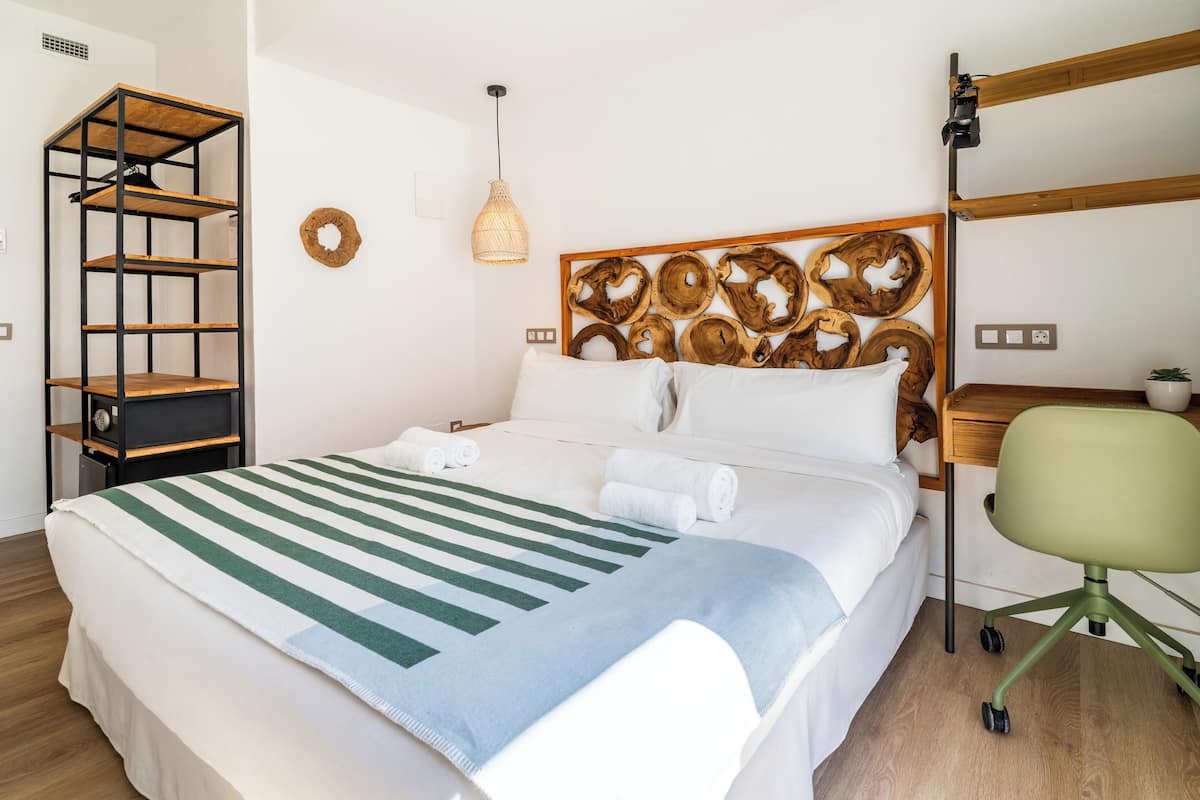 Una cama bien hecha con una manta a rayas, un cabecero de madera con un diseño circular, un estante de mimbre y una silla de oficina verde.