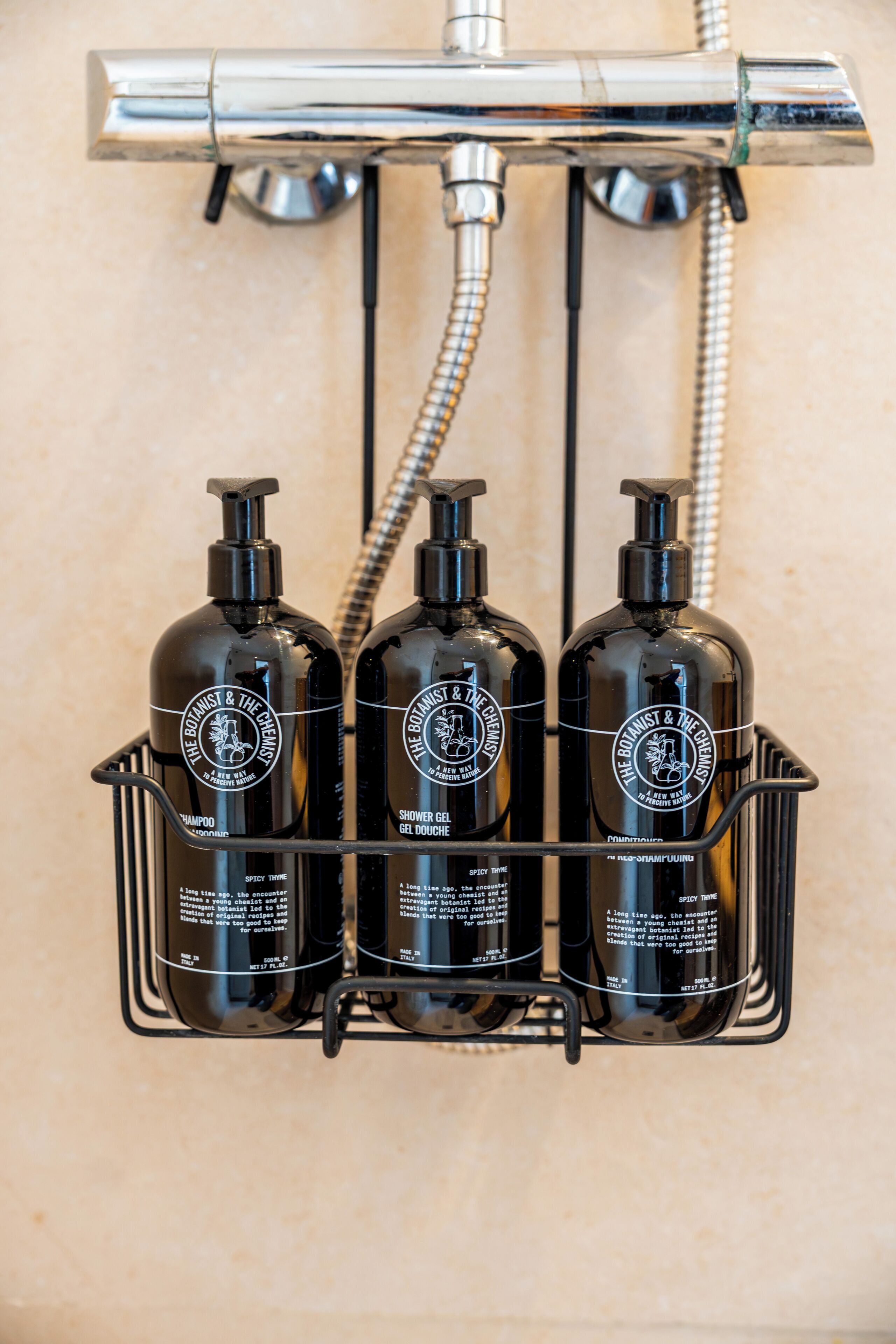 A bathroom with a shower head, three bottles of shampoo and shower gel, and a metal rack.