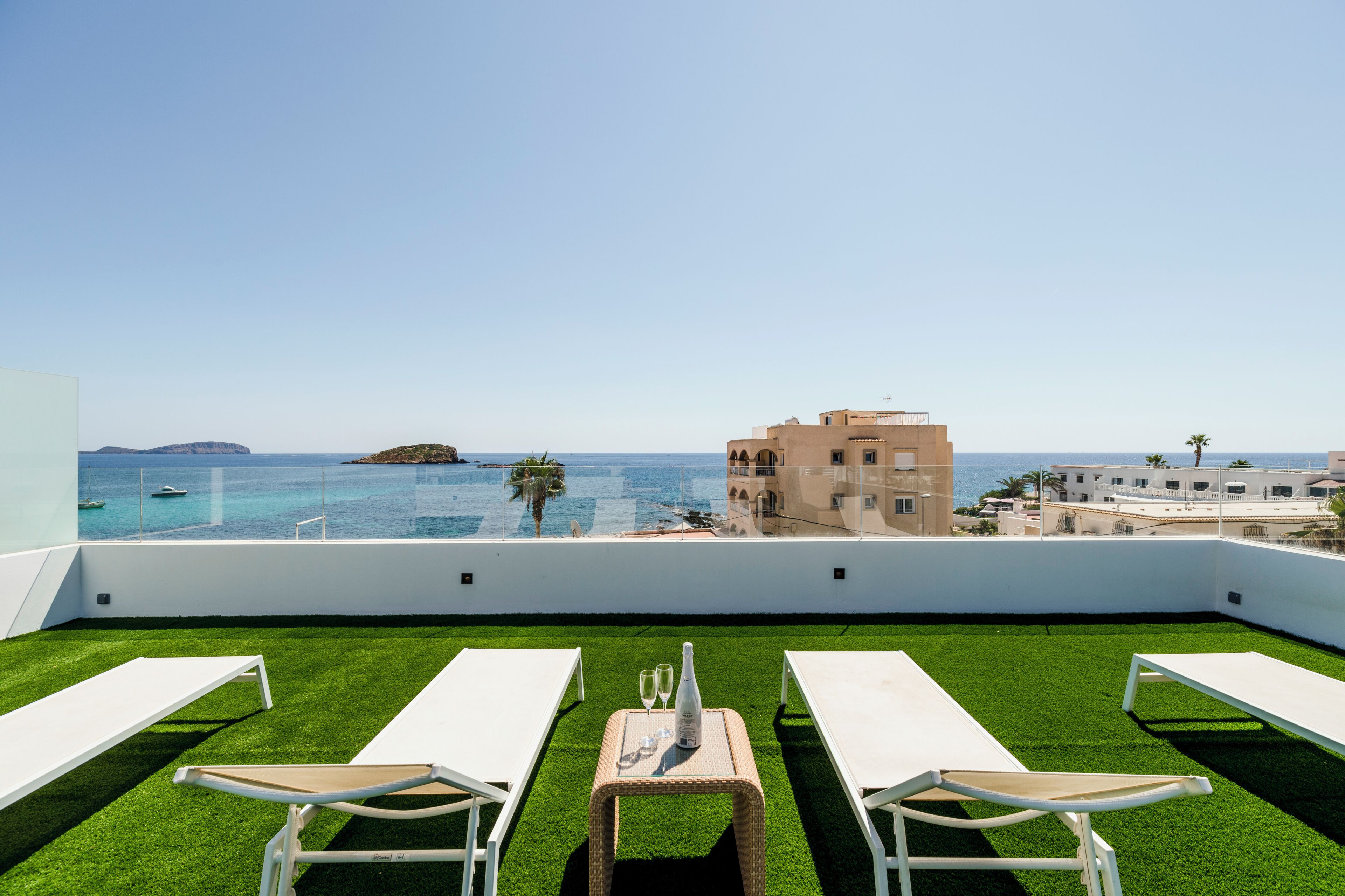 Terraza en la azotea con césped artificial, sillones blancos, una mesa con una botella y vasos, y vistas al mar y edificios.