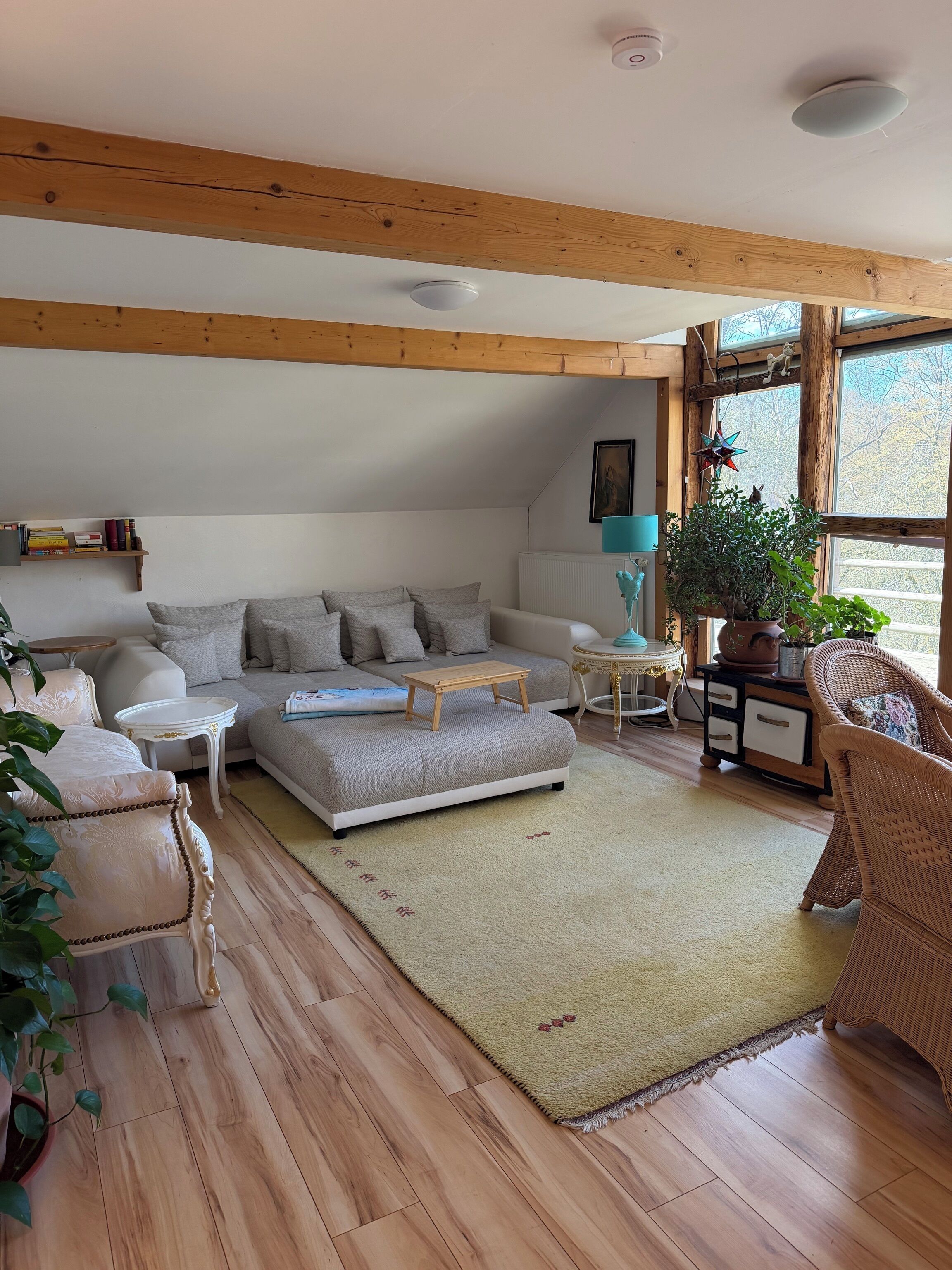 Ein gemütliches Wohnzimmer mit einer Couch, einem Couchtisch und Sesseln, Holzbalken an der Decke und einem großen Fenster.