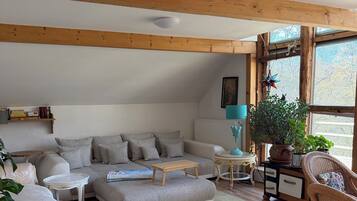 Ein gemütliches Wohnzimmer mit einer Couch, einem Couchtisch und Sesseln, Holzbalken an der Decke und einem großen Fenster.