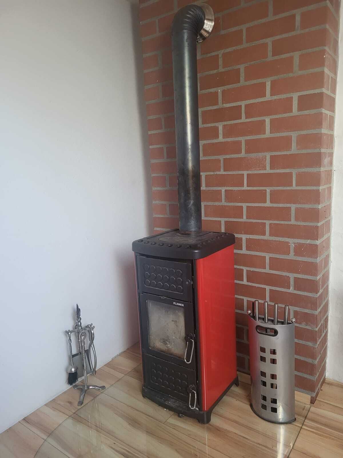 A red and black wood stove with a chimney, a metal brush, and a metal container with tools.