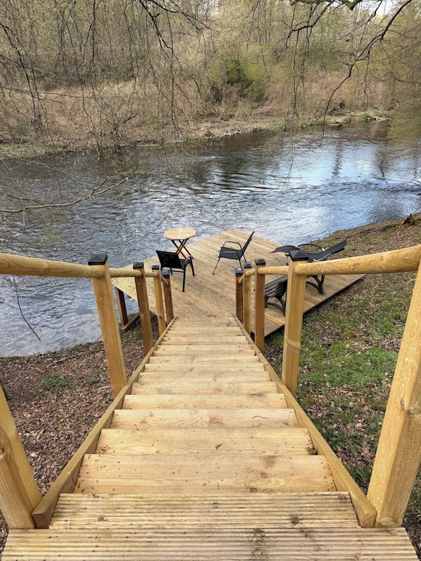 Eine Holztreppe führt zu einem Steg mit Stühlen und einem Tisch an einem Fluss.