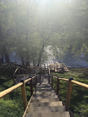 Eine Holztreppe führt zu einer Plattform mit Geländer, umgeben von Bäumen und einem Gewässer.