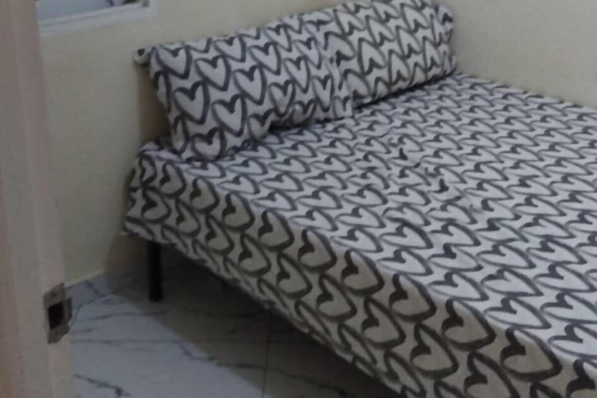 Ein kleines Zimmer mit einem Einzelbett, einer Bettdecke mit Muster, einem Fenster mit heruntergelassener Jalousie und einem Fliesenboden.