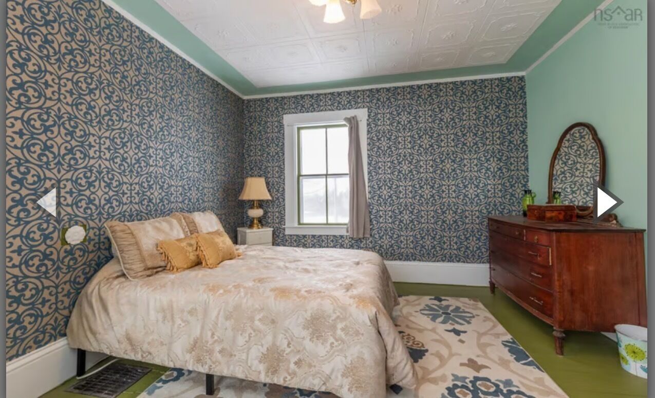 Un dormitorio con papel tapiz estampado, una cama con cubrecama color beige, un aparador y una ventana con cortinas.
