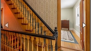 A wooden staircase with a black handrail leading to a room with a patterned carpet and a wooden dresser. - Heart of Pubnico (Pubnico)