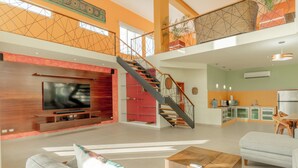 Amplia sala de estar con televisor grande, una escalera y zona de cocina.