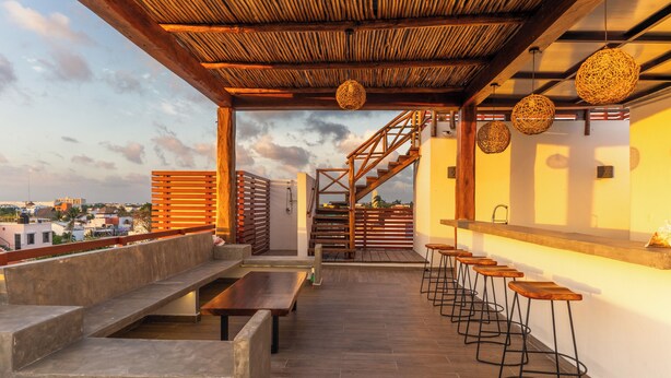 Una terraza en la azotea con bancos de madera, taburetes de bar, una escalera y lámparas colgantes de mimbre.