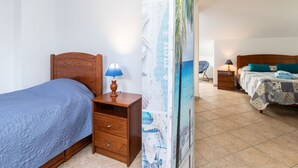 A bedroom with a wooden bed, a nightstand with a lamp, a wall with a beach-themed poster, and a second bed in the background.