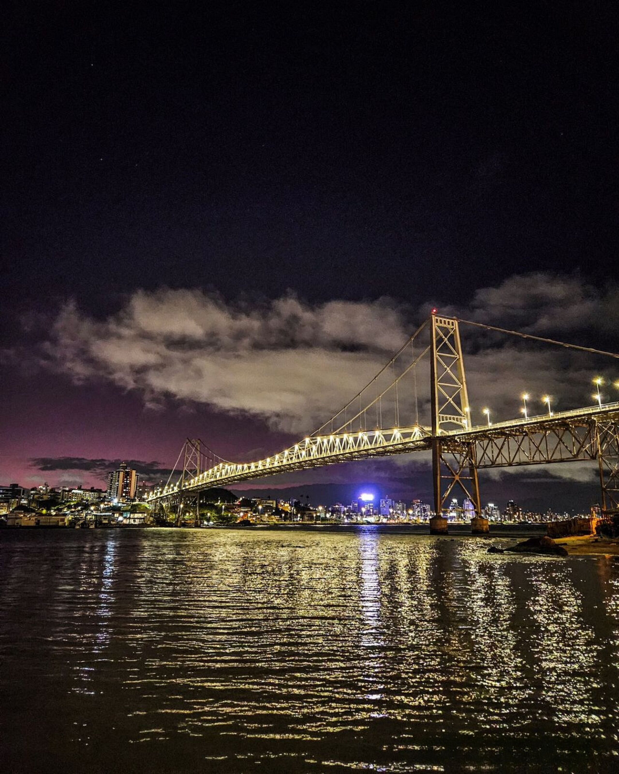 Een nachtzicht op een grote hangbrug met verlichte kabels en torens, overspannend een waterlichaam met lichtjes van de stad die op het oppervlak reflecteren.