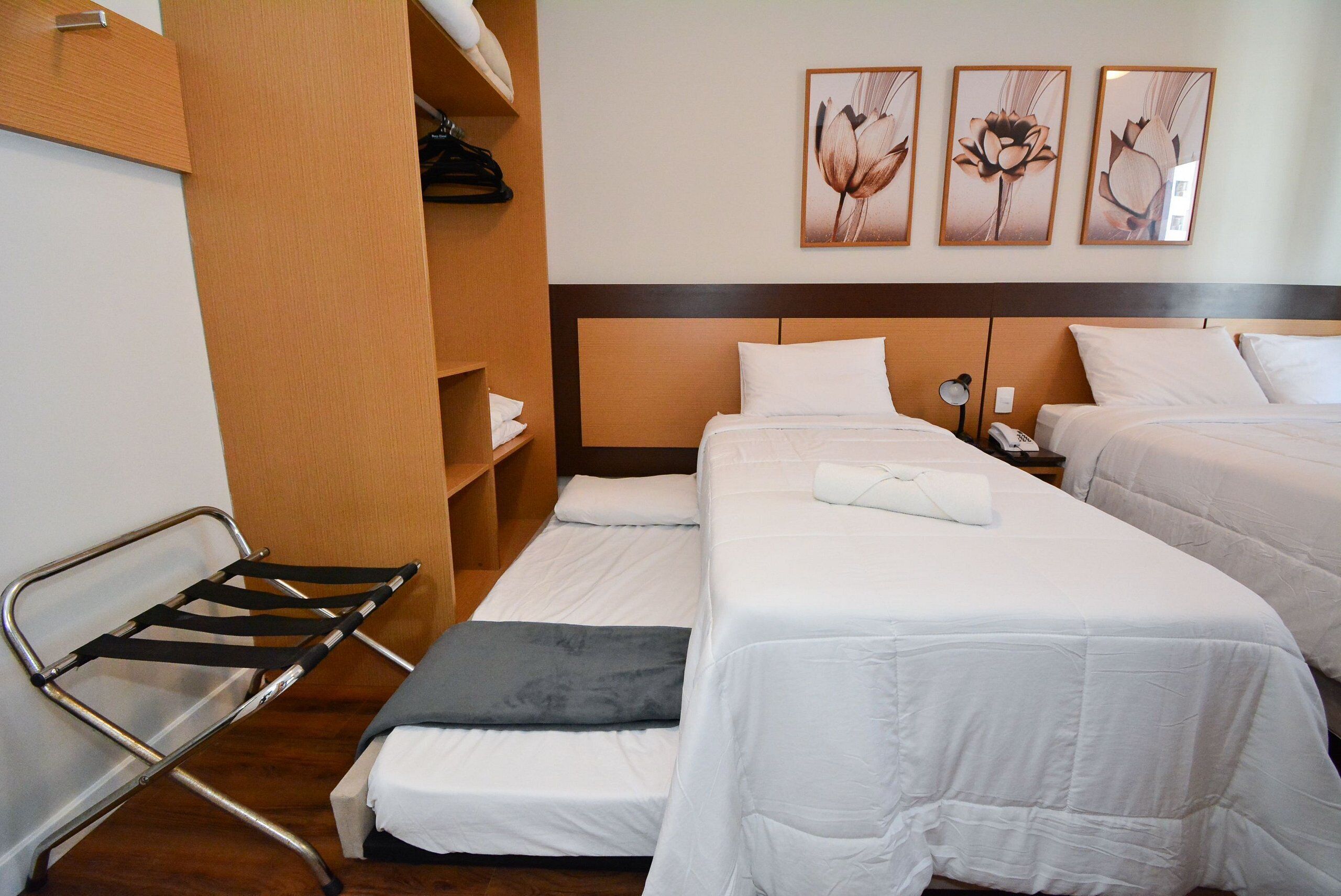A hotel room with two beds, a wooden wardrobe, a metal chair, and three wall-mounted flower paintings.