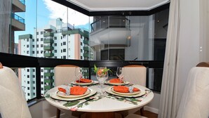 A round dining table with four chairs, set with orange napkins, white plates, and clear glassware, positioned in a corner with a cityscape view through large windows. - Flat no Central Park Residence (Florianopolis)