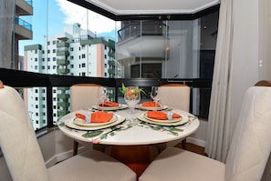 Una mesa de comedor redonda con cuatro sillas, dispuesta con servilletas naranjas, platos blancos y cristalería transparente, ubicada en un rincón con vistas a la ciudad a través de amplias ventanas.