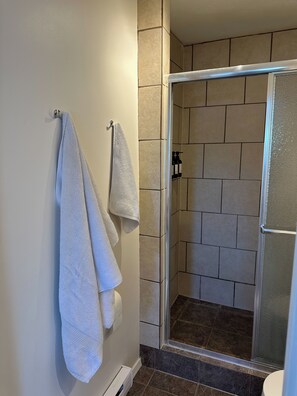 A bathroom with a glass shower enclosure, tiled walls, and a towel rack with two towels. - The Garden Suite (Quesnel)
