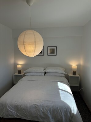 A bedroom with a large bed, two bedside tables with lamps, a hanging light fixture, and two framed pictures on the wall. - The Garden Suite (Quesnel)