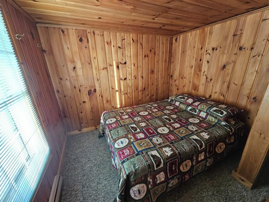 A room with wooden walls and a carpeted floor, featuring a bed with a quilted blanket.