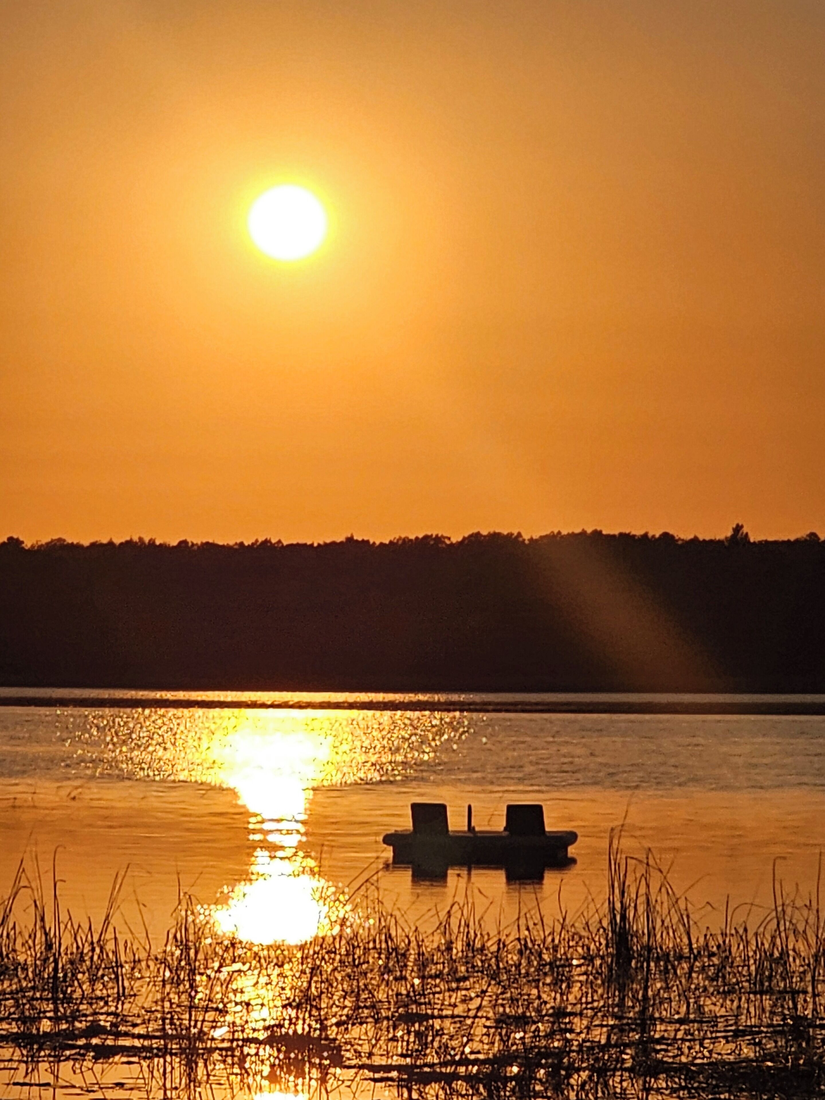 Auringonlasku tyynen järven yllä, etualalla kaksi venettä ja taustalla metsäinen kukkula.