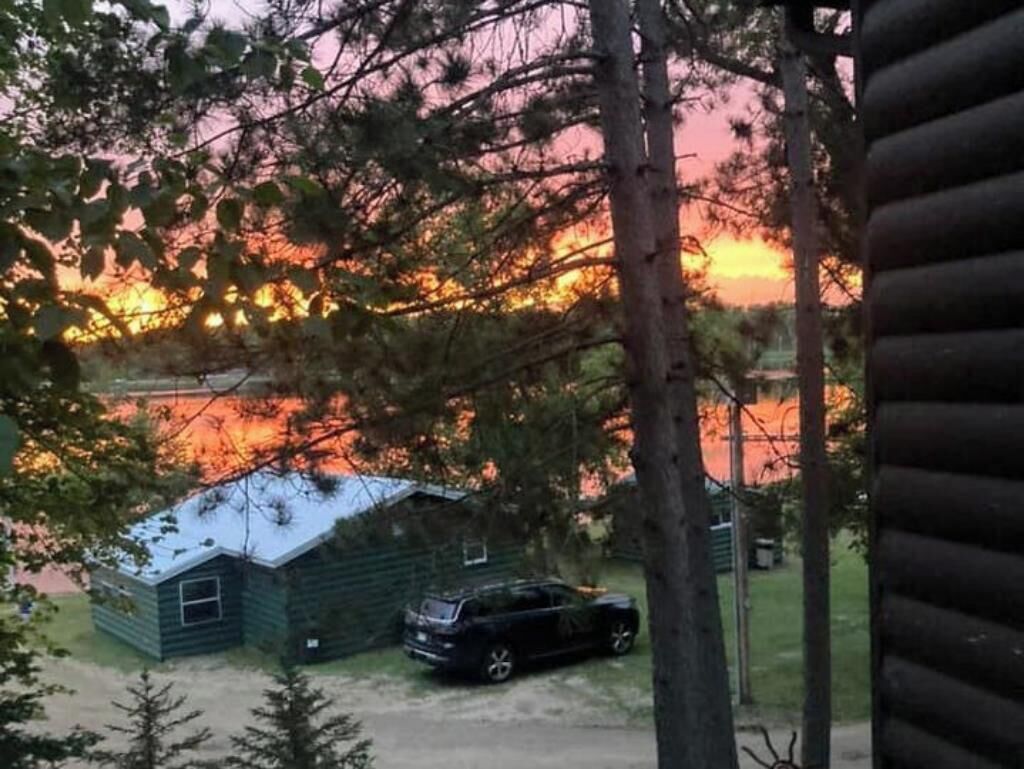 A cabin with a car parked in front, surrounded by trees and a sunset in the background.
