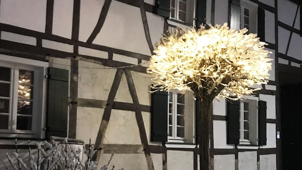 Una casa tradicional con entramado de madera, un jardín cubierto de nieve y un árbol con ramas iluminadas.