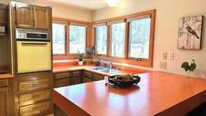 A kitchen with wooden cabinets, a built-in oven, a large island with a bowl, and a window with a view of trees.