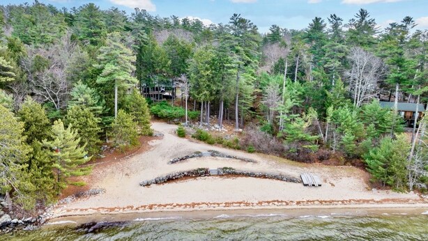 A beach with a sandy area and a small wooden structure, surrounded by dense forest and a building in the background.