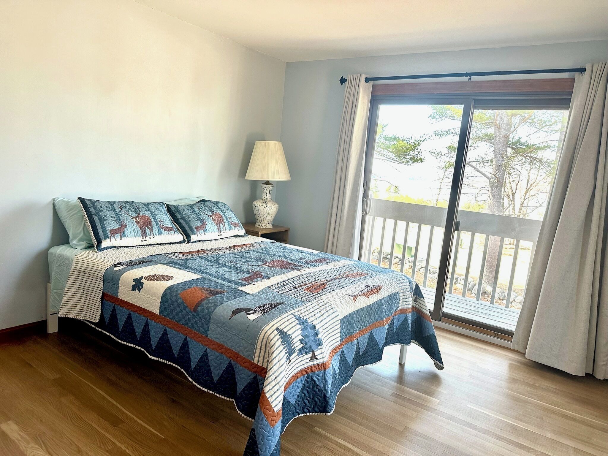 A bedroom with a large bed, a nightstand, a lamp, and a view of the outdoors through a sliding glass door.