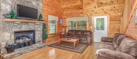 A log cabin interior with a stone fireplace, leather couches, and a ceiling fan.