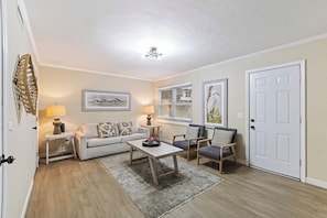 A living room with a sofa, two armchairs, a coffee table, a lamp, a painting, a window with blinds, and a door.